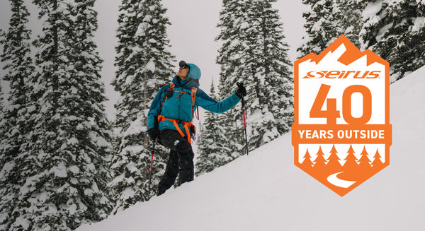 person looking up while split boarding up a mountainside with trees in the background and seirus 40th anniversary logo overlay
