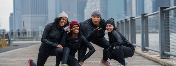four woman wearing base layer with a city backdrop bending down and posing for camera